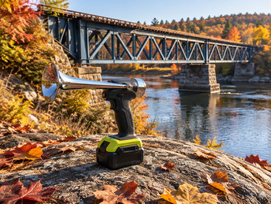 A chrome train horn gun with a green battery sits on a rock beside a steel rail bridge over a river in autumn.