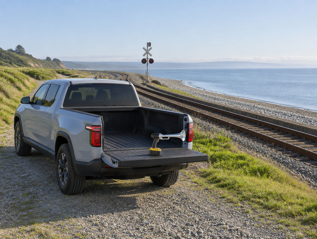 A grey electric pickup parked by a seaside railway track, with a battery horn gun resting on its lowered tailgate.
