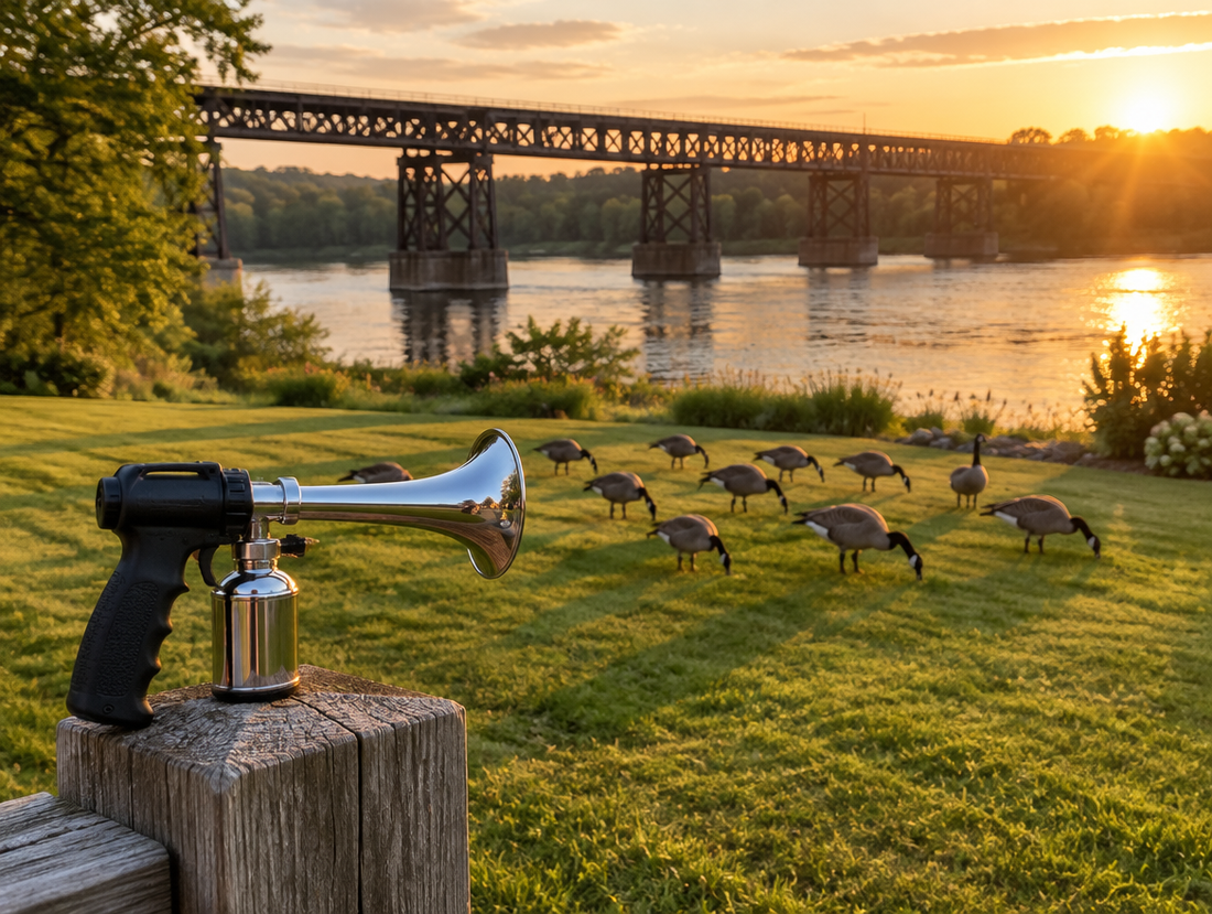 A chrome air horn device on a fence post near geese grazing on a riverside lawn below a steel trestle bridge at sunset.